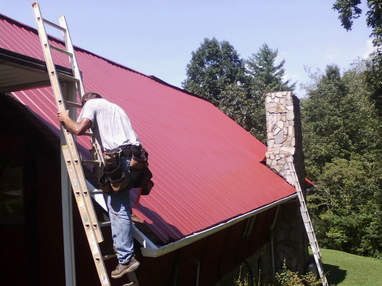 Roofer on a ladder beside a red metal roof and stone chimney — Soffit & Fascia Repair by Roofing Brothers in Rolesville